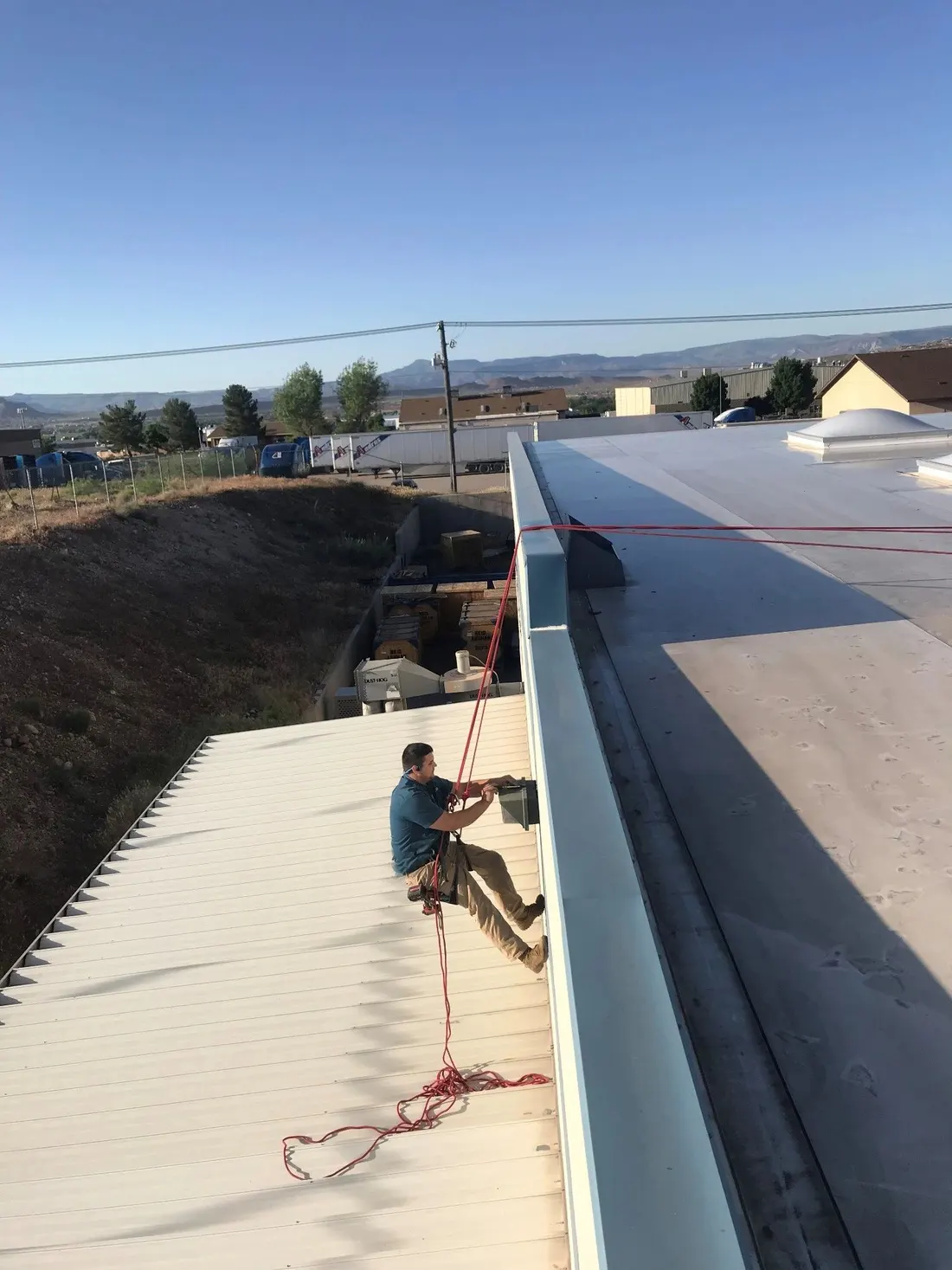 Electrician on a rope line working on a sloped metal roof above a commercial yard